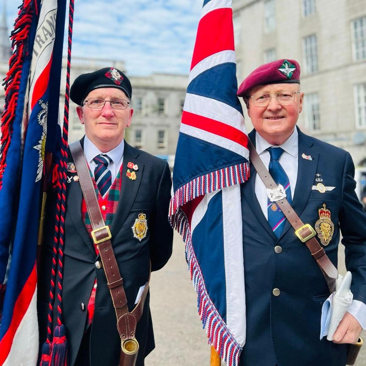 Two men face the camera wearing suits and military regalia carrying large flags