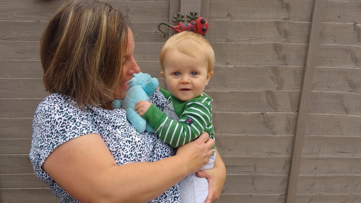 Mum Deborah, wearing a leopard print top, holds her one-year-old son Joe in her arms, as he holds his blue teddy.   