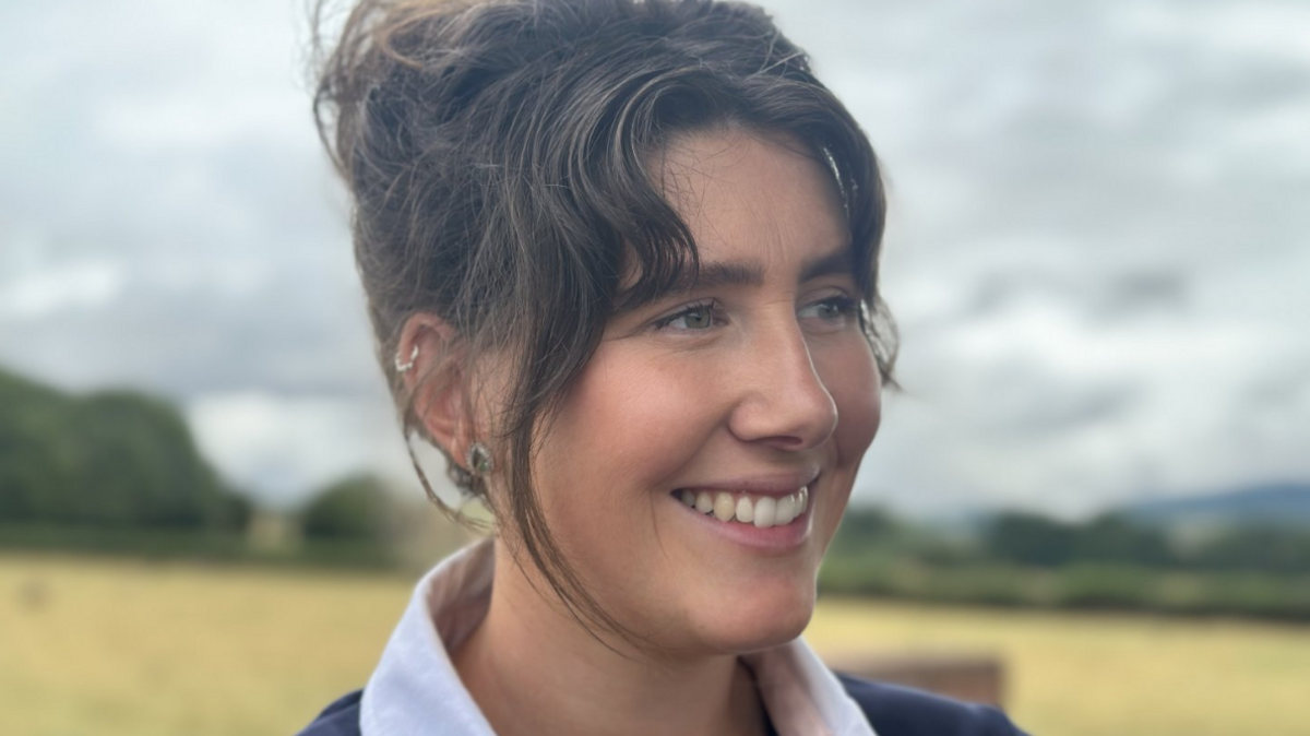 A woman with brown hair pulled into a bun smiles to the side of the camera. She wears silver earrings. The photo is a close-up so you can see her head and shoulders. The background is blurred but you can see the yellow crops on a farm and trees behind her.