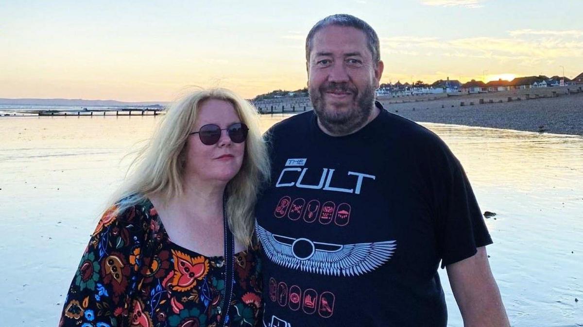 The image shows a couple posing for a picture: the woman on the left has long blonde hair and is wearing a colourful patterned dress with sunglasses. The man on the right has short greyish-brownish hair and a short beard. He is wearing a black T-shirt with the writing "The Cult" on it in white lettering above some symbols.
Behind them is a coast, and the shore can be seen on the right-hands-side with a few houses and the Sun peaking behind them.