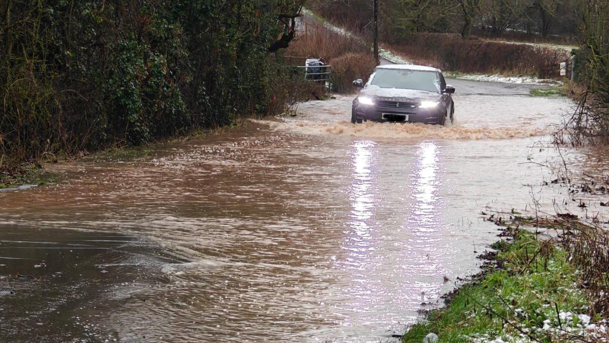  A vehicle travelling through flood water, with hedgerows visible either side of the road.