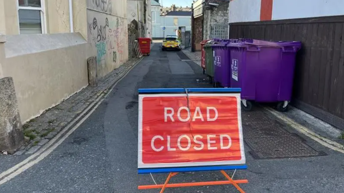 A red sign with the words 'road closed' on it in the middle of a narrow road in Mutley. There are two large purple bins on the right, next to a fence, and a police car at the end of the road in the distance.
