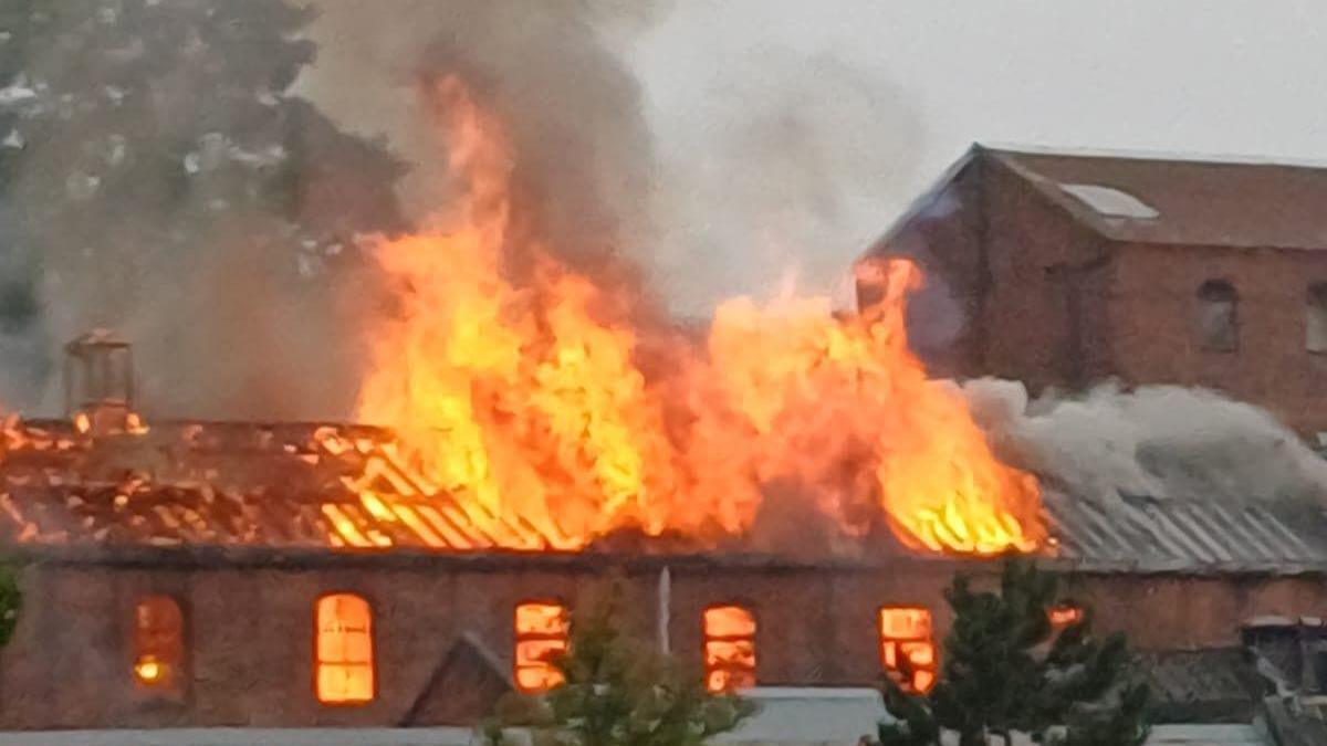 Large orange flames can be seen through the roof of the large brick building.