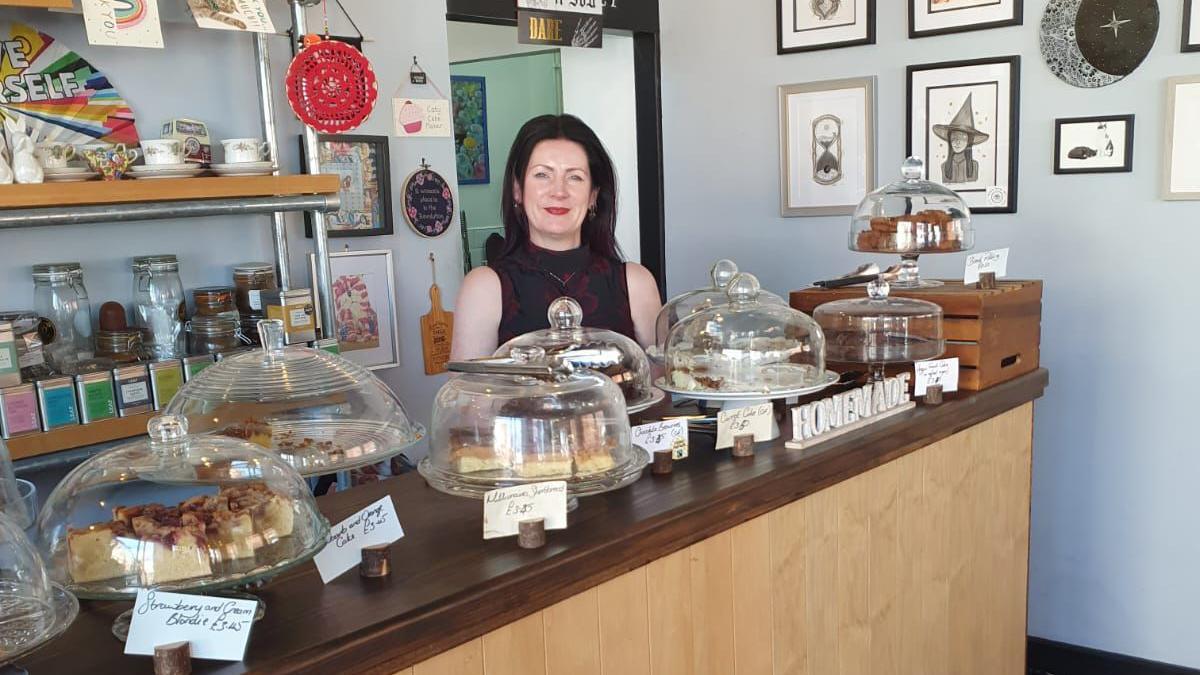 A woman stands behind the counter of a cafe. She has straight black hair and she is wearing a black and red sleeveless top. The counter has several cakes on it, under domed glass lids.
