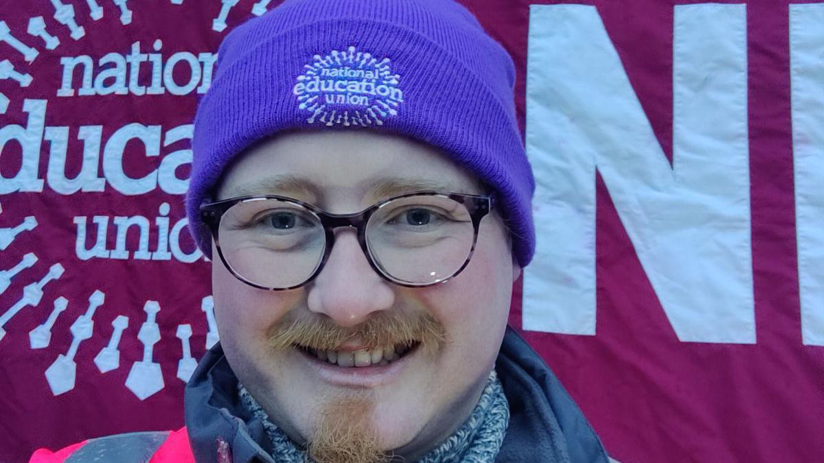 A man is seen smiling directly into the camera. He is wearing a pink hi-vis jacket, and has a purple hat reading National Education Union on it. He has glasses on, and distinctive facial hair.