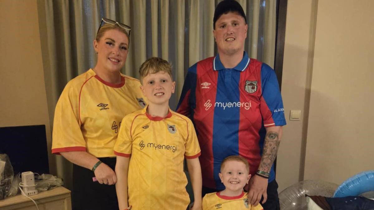 A family comprising a man, woman and two boys are smiling and wearing Grimsby Town shirts. They are standing in their hotel room in Turkey.