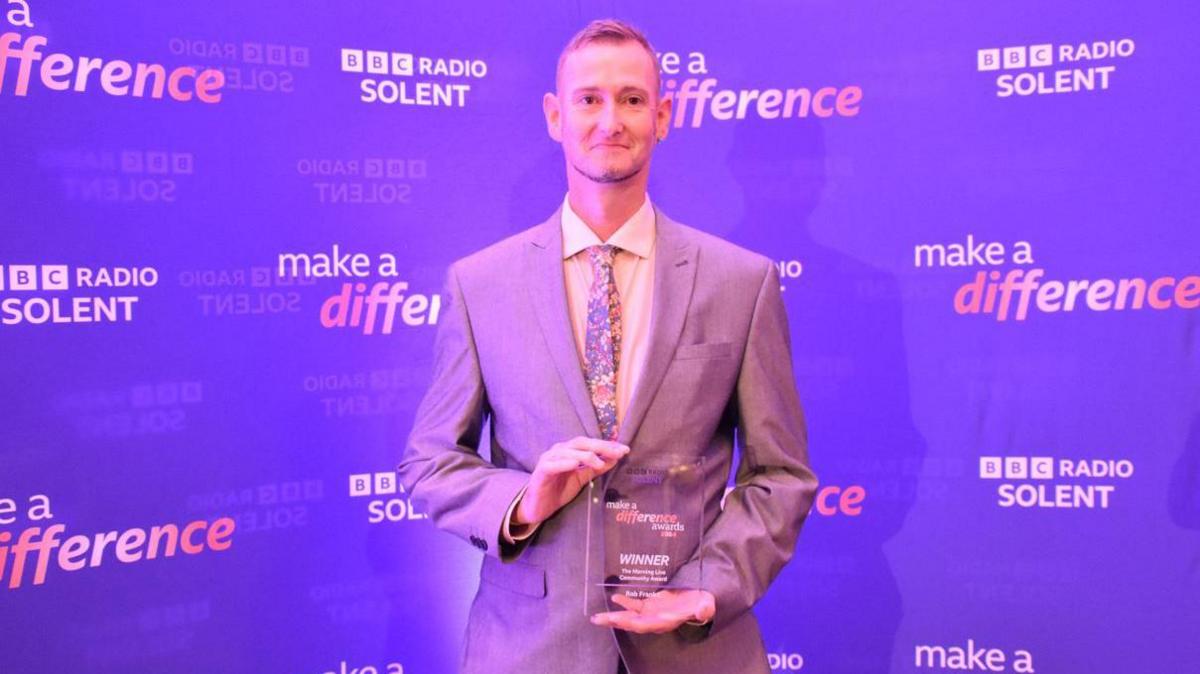 Rob Franks holding his award, wearing grey suit, in front of a purple wall, reading "BBC Radio Solent make a difference"