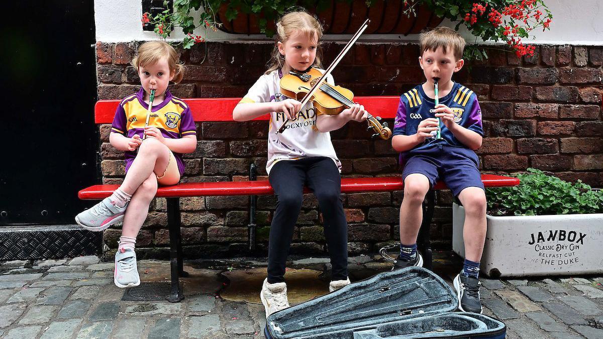 Three children are sitting on a red painted wooden bench on a cobbled street. A boy and girl are playing the tin whistle and another girl is playing the violin. 