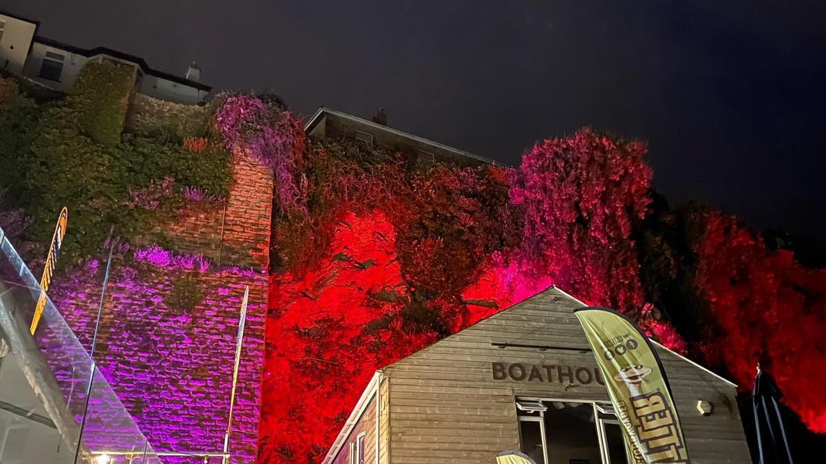 The ivy-covered wall of Newquay Harbour is lit up with red, purple and pink lighting. In the foreground is a building with the letters BOATHOUSE on it and a flag advertising Cornish Rattler.