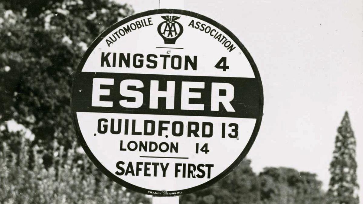 A black-and-white image of a circular road sign for Esher, showing distances to Kingston, Guildford and London as well as the Automobile Association logo and the message "Safety First"