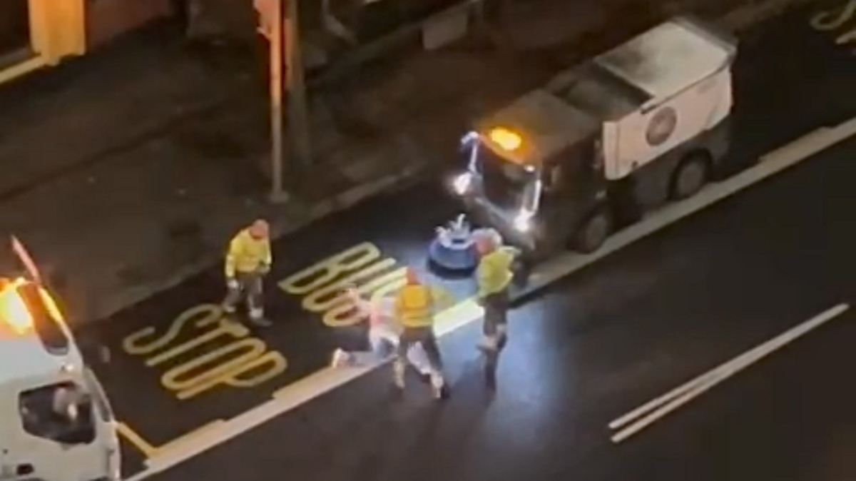 Three men in high vis jacket are standing around a man lying on the road. Behind them there is a Belfast City Council van.