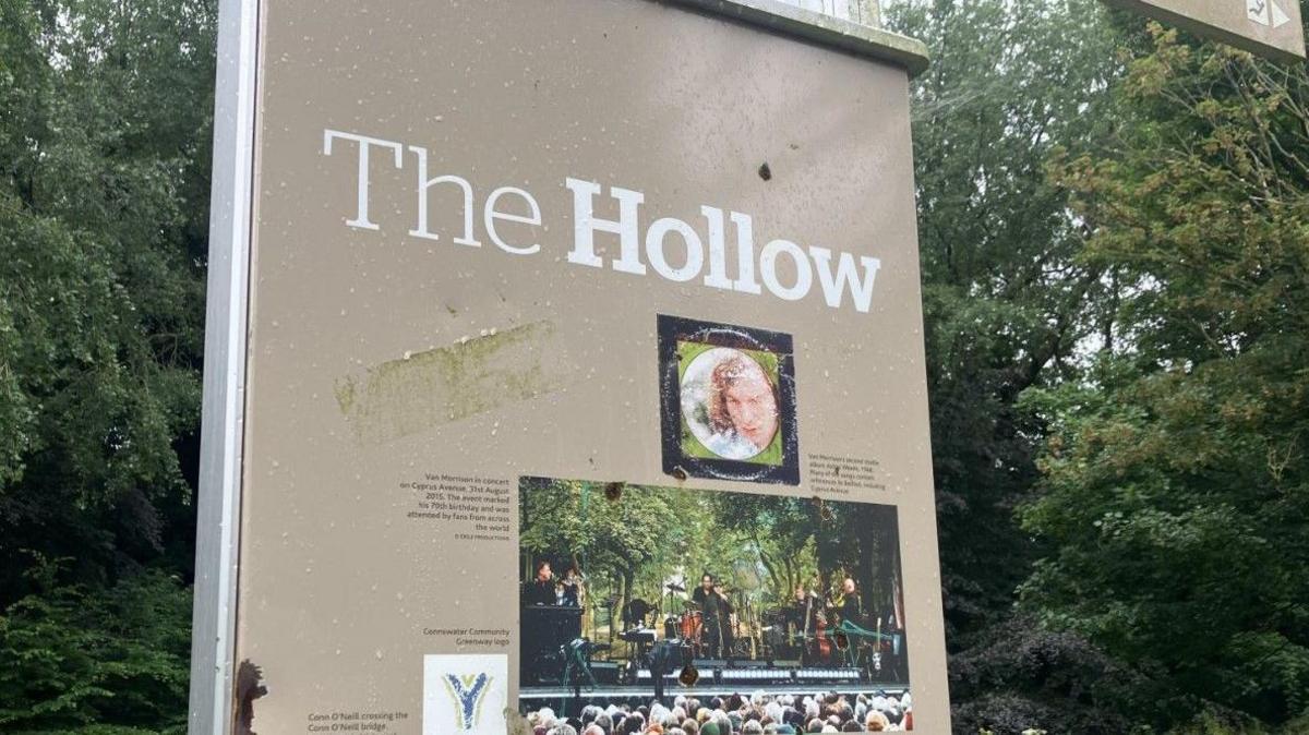 A tall beige upright sign which has the words 'The Hollow' at the top. There are small images of Van Morrison, one from the cover of his album Astral Weeks, and the other taken from a live performance. Behind the sign are lush green trees. 