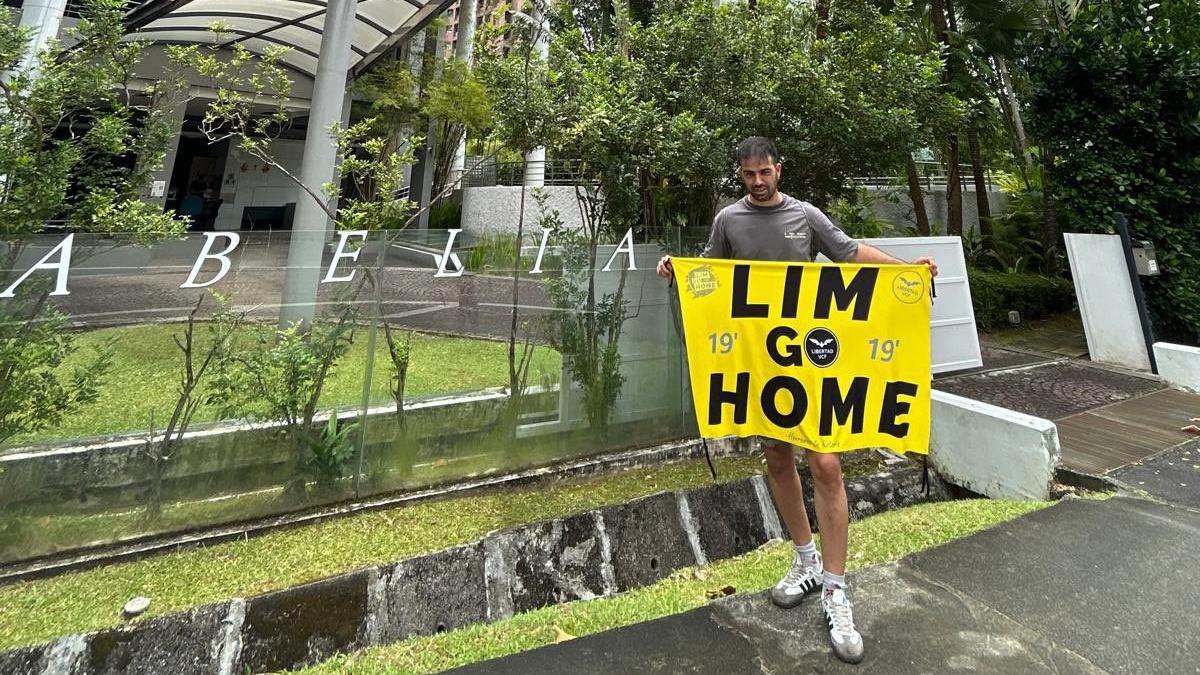 A man outside a residential apartment complex holding a yellow banner that reads LIM GO HOME