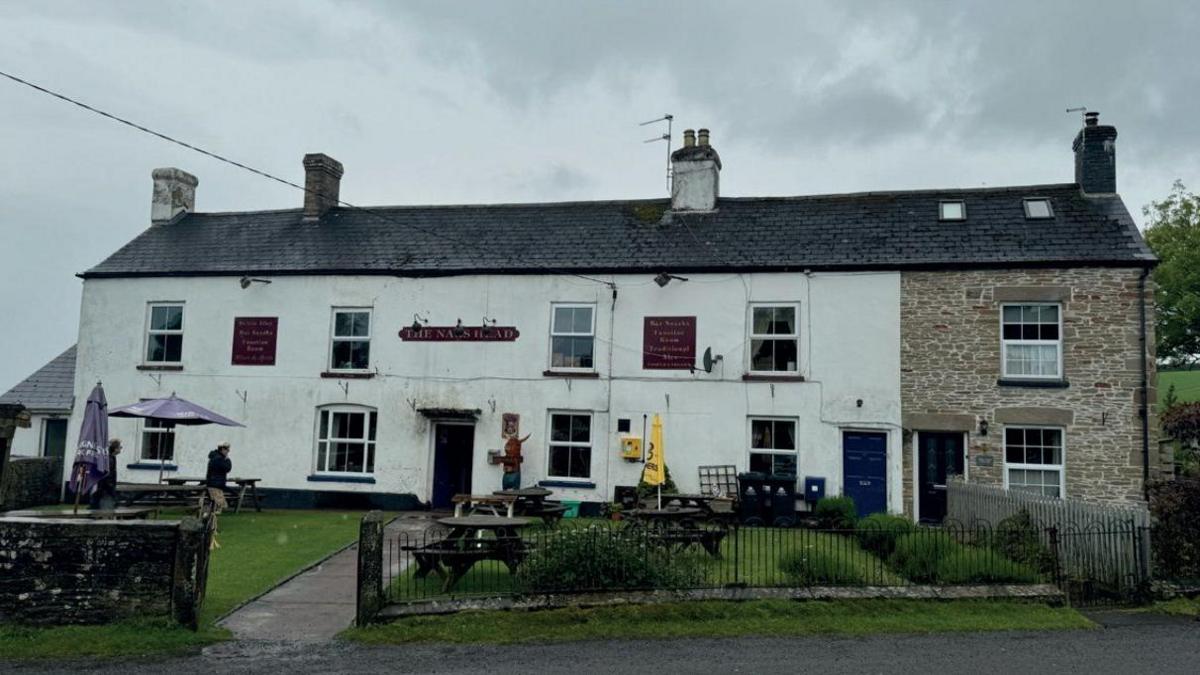 Two storey pub, with four well-spaced windows on each floor. There is a grass garden in front with round benches and a black metal fence. A small red and gold sign above the door reads The Nag's Head