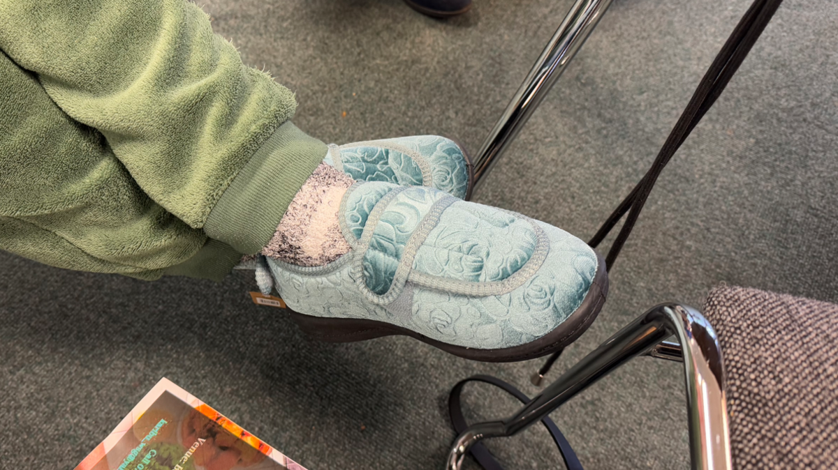 A pair of light blue coloured slippers on the feet of an elderly woman wearing green jogging bottoms.