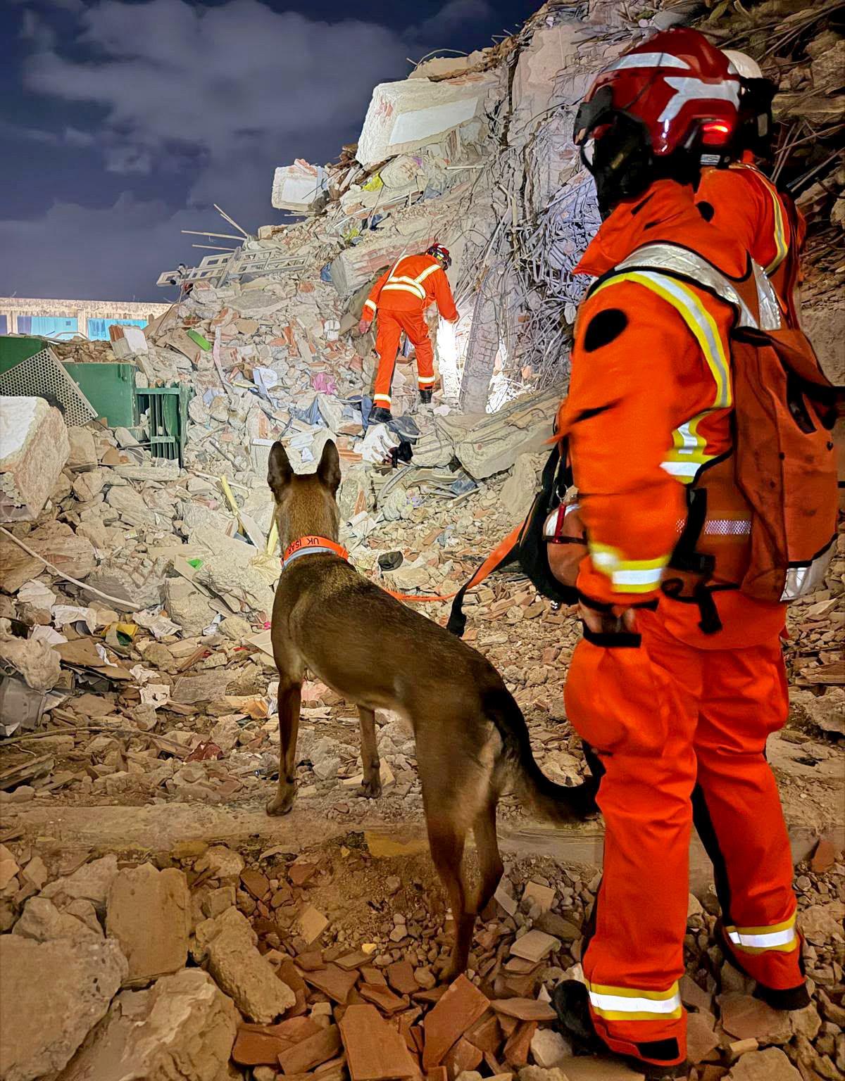 A person dressed in head-tot-toe orange hi-vis holds a dog on a lead, as they look for survivors in an area covered in rubble in La Guaira, Venezuela.