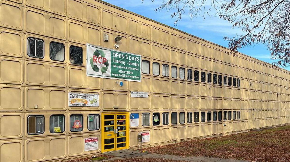 The outside of a warehouse in Milton Keynes where the MK Reuse charity is based. Outside is grass covered in autumn leaves and the branches of trees. The building looks like a pre-fab warehouse, it is a cream colour and is covered in panels with moulded squares. There are bright yellow doors and posters around the doors.