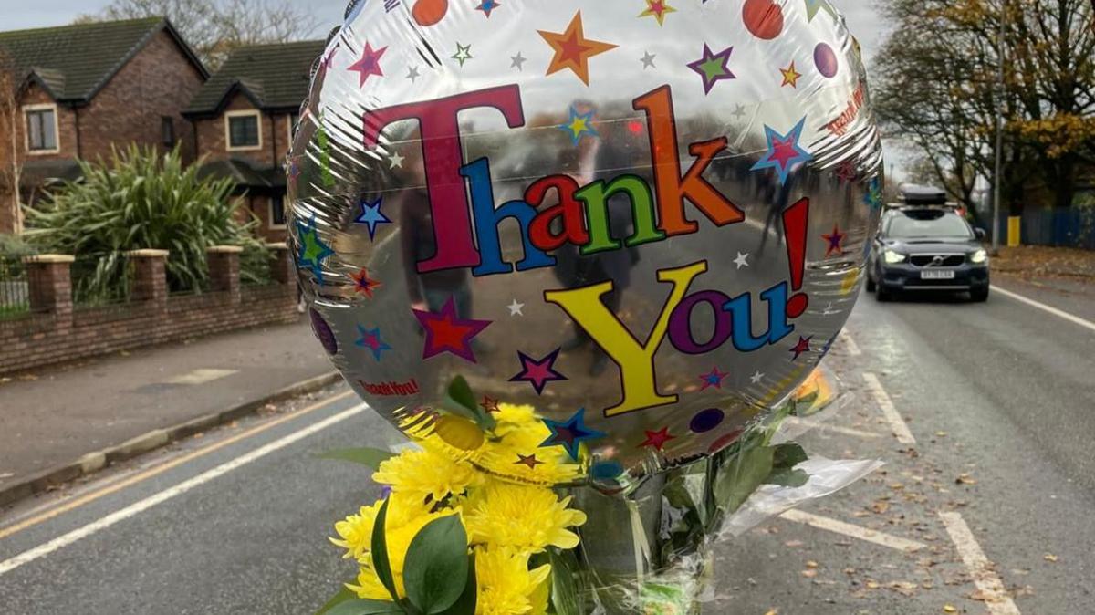 A silver balloon that reads 'Thank You' in multi-colour text is tied to a lamppost on a central reservation. A bunch of yellow has been tied to the lamppost.