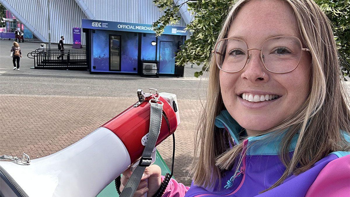 Alice McNaugher, sitting in a tall chair similar to those used by tennis umpires. She is wearing a purple and pink jacket and holding a megaphone in one hand. She is sitting outside at the SEC campus in Glasgow.