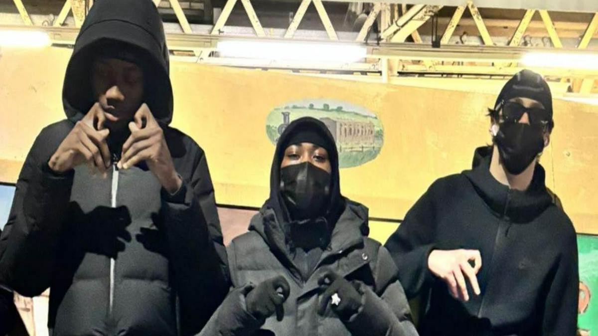 A photograph of Camron Osei, Shiloh Hinds and Bradley Harris who were all dressed in dark clothing, with masks and hoods. The trio are posing next to a graffitied sign next to Harrow and Wealdstone Tube station.