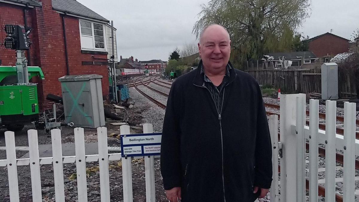 Dennis Fancett stands at Bedlington station with a white fence behind him and two tracks merging behind him. There is a small signal box behind him. 