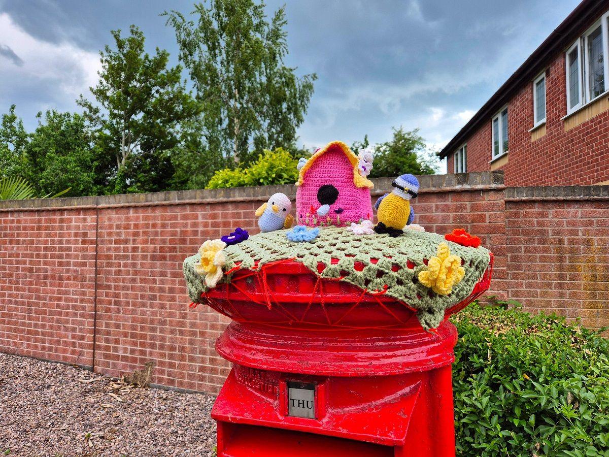 Woollen figures including a couple of birds and a bird box, knitted from wood, adorn the top of a red postbox which is in front of a brick wall.