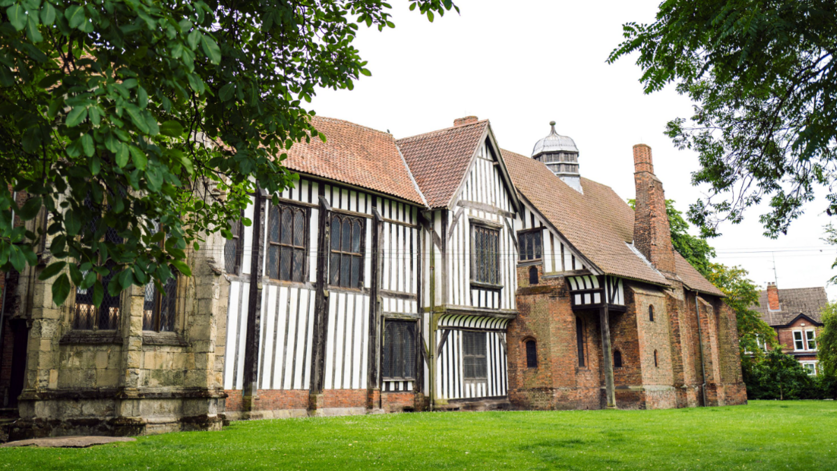 An outside view of Gainsborough Old Hall. It has a white and black exterior as well as orange and beige brick. Trees cascade over the top of the picture.