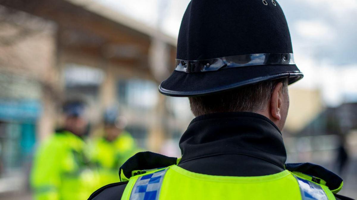 A picture of a police officer taken from behind. The male officer is where his hi-vis jacket and his police hat.