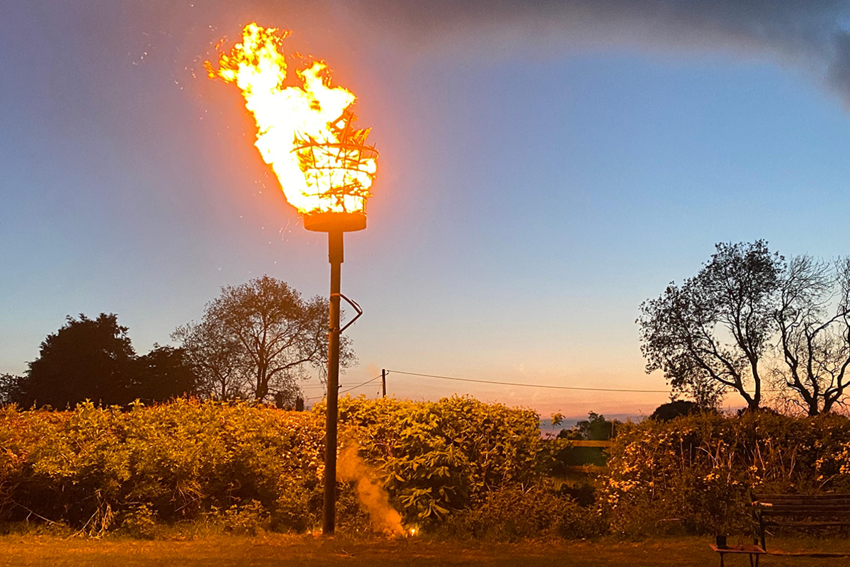 A blazing brazier on a tall pole in a field at twilight, with a hedge in the background