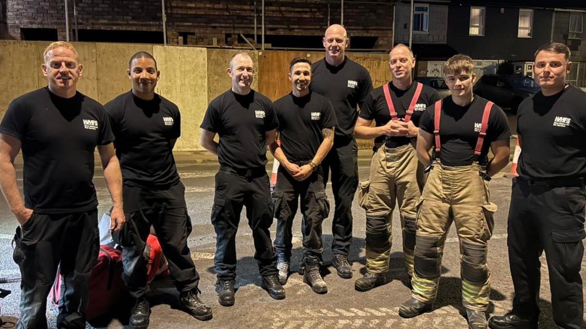 Eight men are wearing black t-shirts and trousers with the logo 'West Midlands Fire Service'. They are standing on a street and smiling.