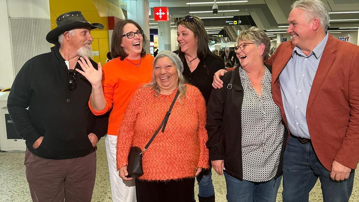 Six people grouped together at an airport smiling and laughing. Tina wearing a bright orange jumper and doing jazz hands.