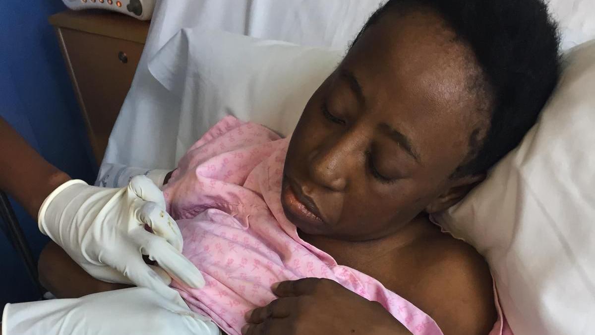 The gloved hands of a nurse lift up a pink hospital gown worn by Toks Odesanmi. She has just woken up after her transplant and looks tired.