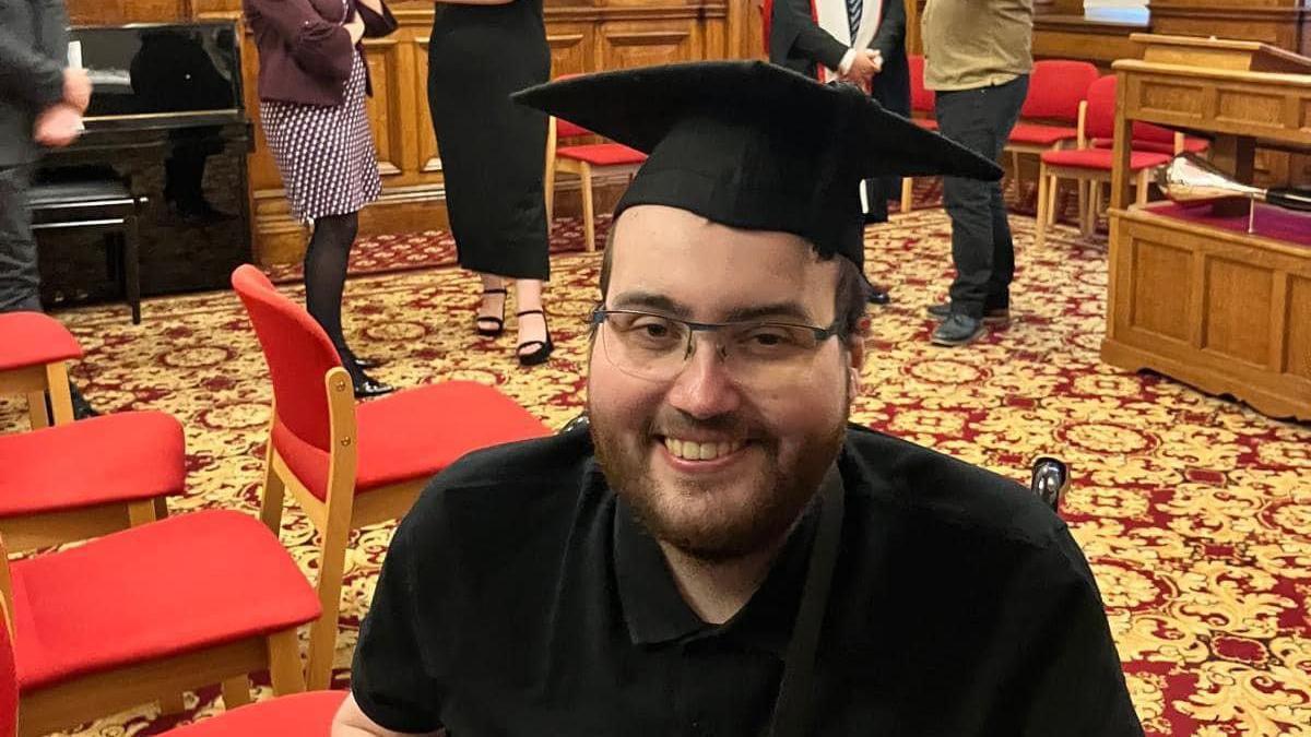 Daniel Swales, in a black t-shirt and with a black graduation cap on, is smiling at the camera. He has brown beard and is in a hall with red chairs.