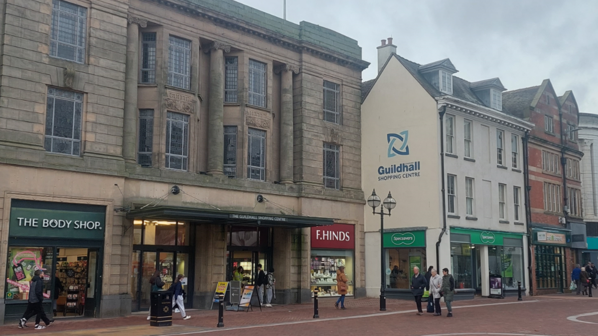 A row of grey shops with signs reading The Body Shop and F Hinds on the ground floor. There is a sign reading Guildhall Shopping centre half-way up one wall. A few people mill around in front of the stores.