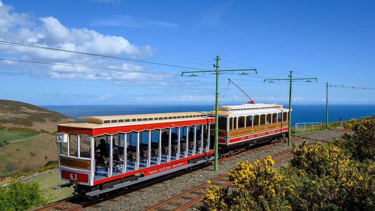 Two electric trams sit on tracks on a cliff top, you can see green bushes with yellow flowers in the foreground and blue skies and sea in the background.