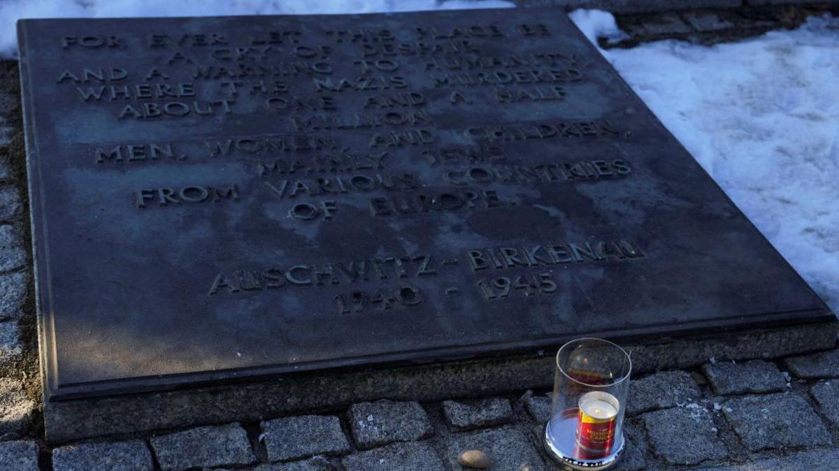 A candle in a clear jar burns beside a plaque marking the liberation of the Nazi's largest concentration camp at Auschwitz-Birkenau. it is surrounded by snow.