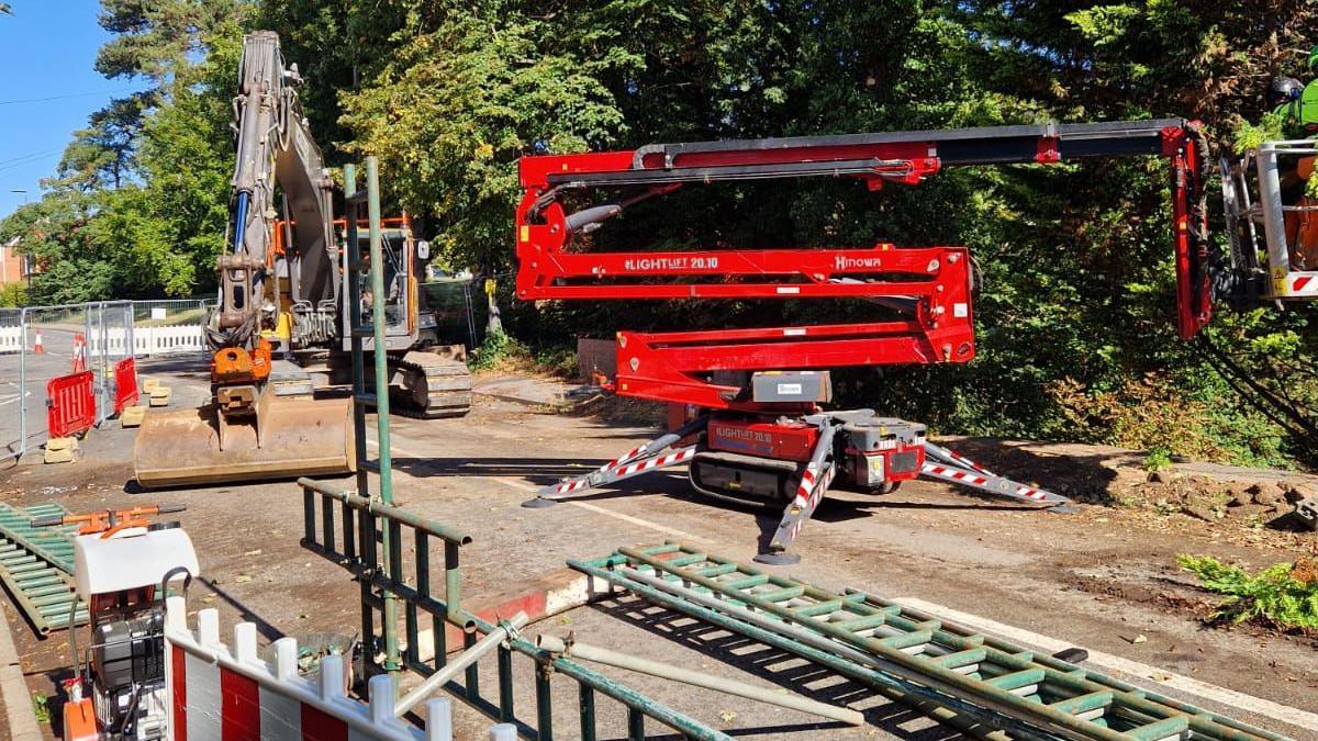 A photo showing a red crane folded down and an orange digger on a building site. There is various equipment in shot with some temporary fencing around the diggers. There are trees in the background.