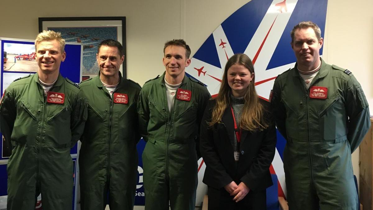 Holly stands in line with four Red Arrow pilots. The men were green pilot suits. Holly has long brown hair that is down and she wears a black suit.