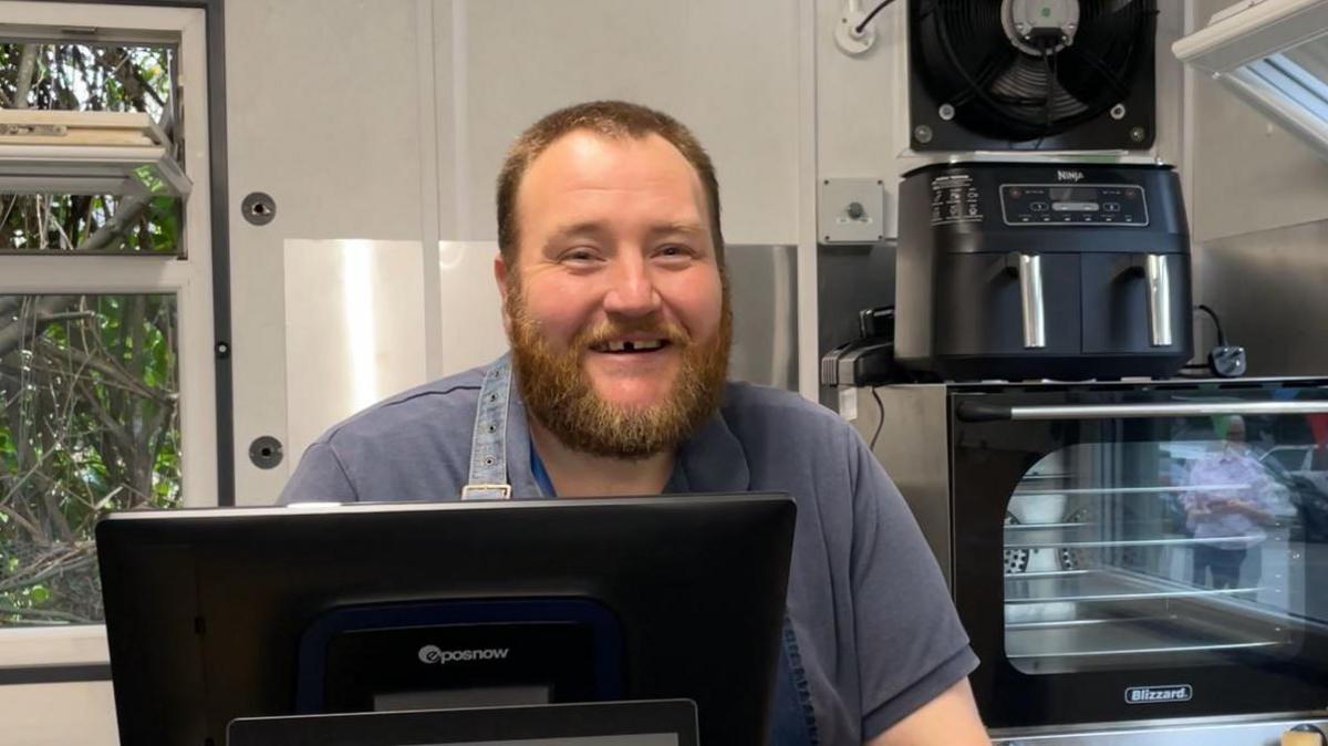 Dan stood behind the counter of the shop. ovens and pots are behind him. He is wearing a grey t shirt and a dark colourd apron. He has a ginger beard and short hair. 