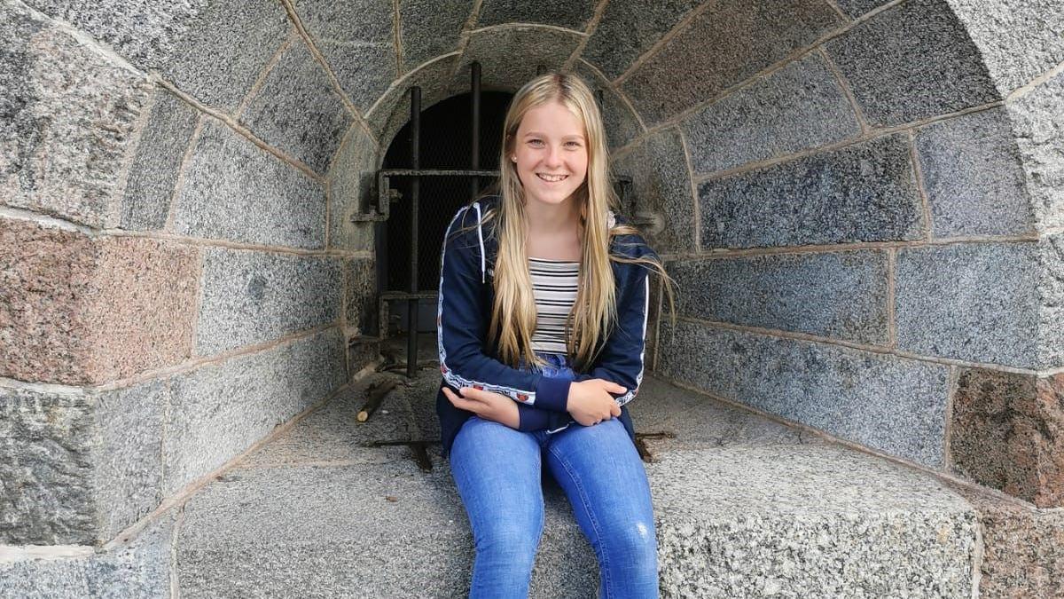 Gaby is sitting on a stone ledge as a young girl. She has long blonde hair and is smiling while wearing jeans and a stripy top.