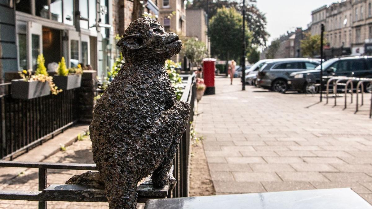 A metal statue of a Rhesus monkey perched on a railing on the side of a pavement in Clifton. It is looking upwards, as if watching people walk by. The metal has a stippling effect to it to emulate fur.