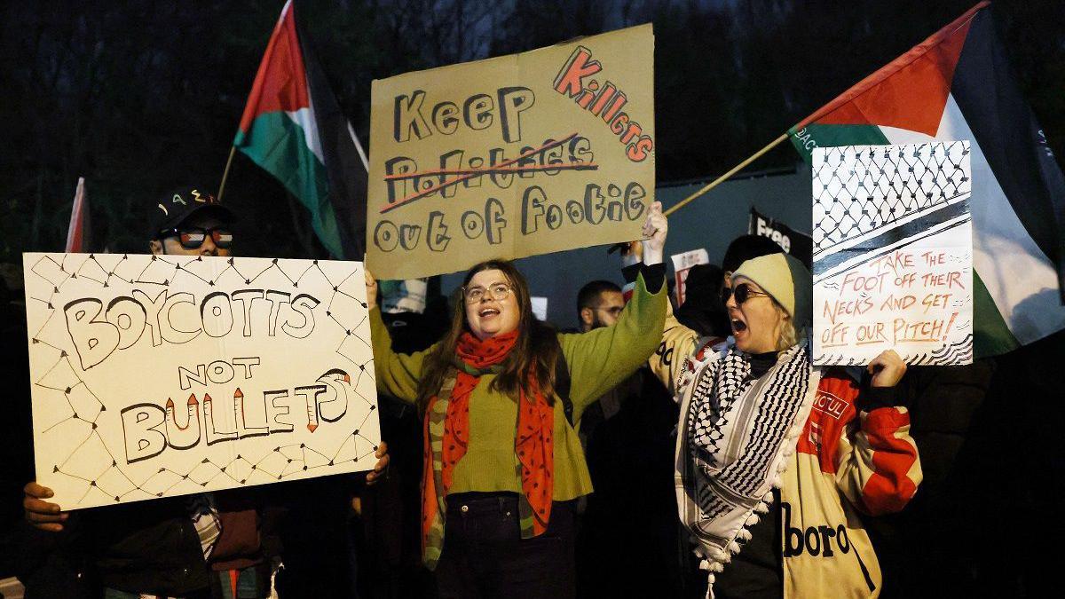 Protesters hold signs outside the stadium before the match. They have Palestinian flags. Two signs say "boycotts not bullets" and "take the foot off their necks and get off our pitch".