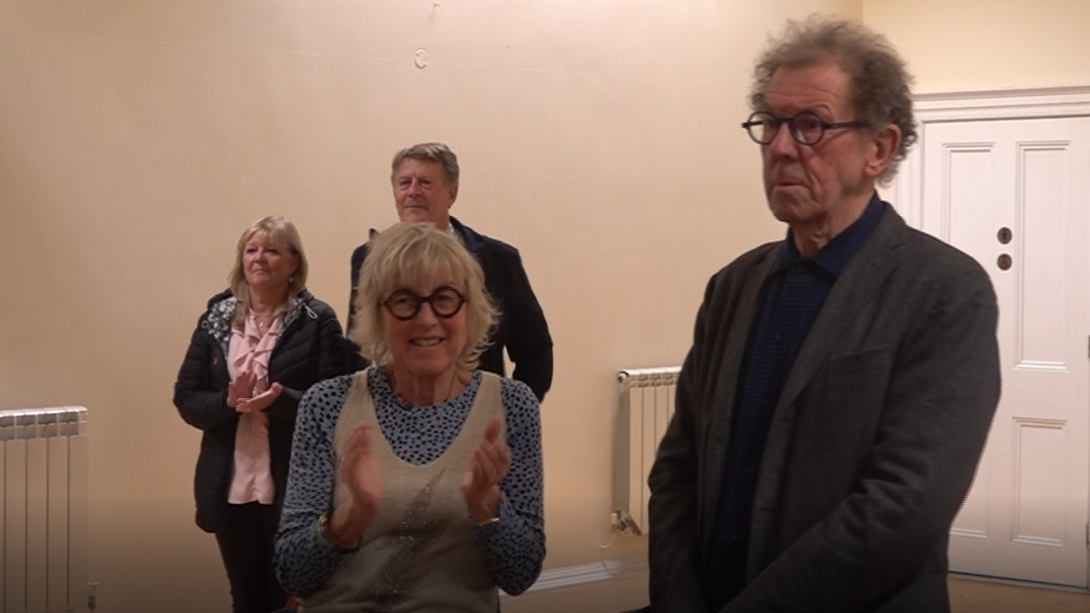 William Tate stood to the right wearing a grey blazer and dark blue shirt. He is wearing black glasses. To the right is a woman wearing a light blue top with a vest over the top. She is wearing black glasses and is clapping. There are also two people stood behind them, on the left, who are also clapping and smiling. 