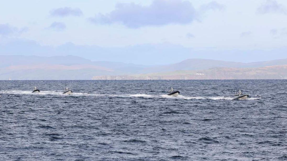 Four of the white and black Rattlers sail through the water, leaving behind white waves. In the far distance are green mountains.