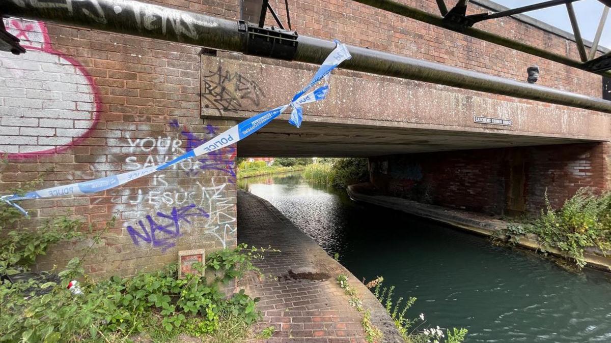 Police tape hangs from a metal bar in front of a bridge over a canal. Several pieces of graffiti have been painted on the bridge.