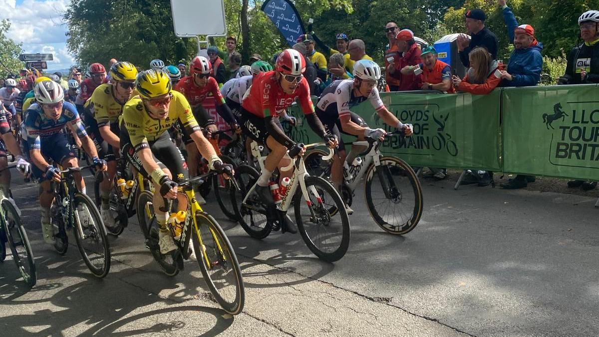 A cluster of cyclists wearing a mix of blue, yellow, red and white outfits speed past spectators behind a green barrier.