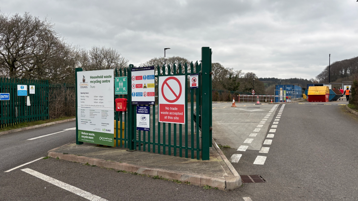 The entrance of Truro's household waste recycling centre. The sky is grey and cloudy in the background and there are workers in the distance on the right.