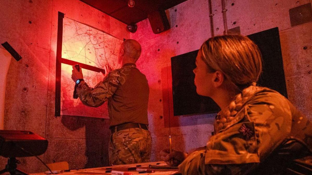 An immersive room that is used to replicate front lines being used in Warminster. Two military personnel are looking at maps in a room using a red light.