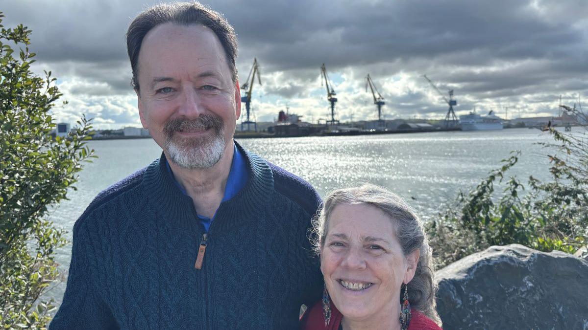 Randy and Kit standing in front of Belfast's ship yard. Randy is wearing a navy zip-up fleece with a blue top and Kit is wearing a red cardigan with a black top underneath.