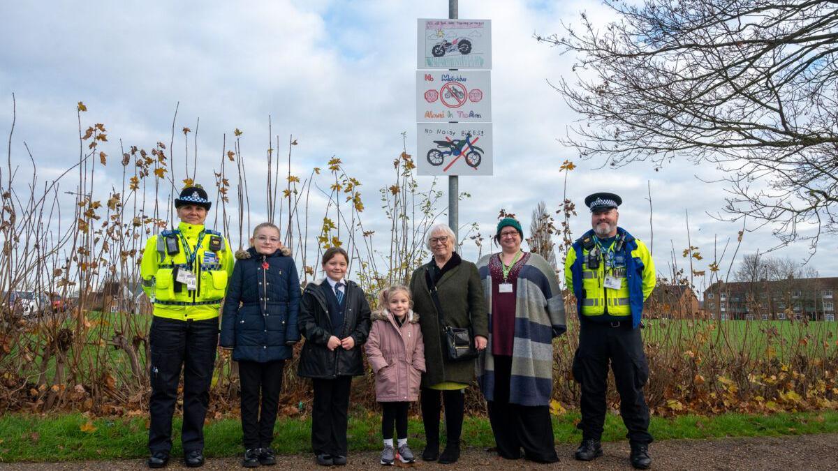 Two police officers in hi-vis jackets stand next to three children, a woman in an olive coat with glasses, and a woman in a grey, navy and grey striped top. They are standing in front of a metal pole with the anti-nuisance motorbike signs